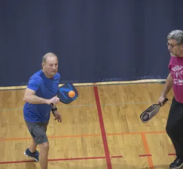A pickleball game at the Ragsdale YMCA. Two men on a team stand together. The man on the left is sending a bright orange ball back to their opponent.