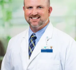 A portrait of Dr. Mark Cairns, MD, MS. A man wearing a white laboratory coat smiles at the camera. He is wearing a blue striped button down shirt with a dark blue tie with yellow and light blue stripes.