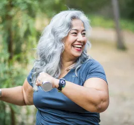 A senior woman smiles while exercising using hand weights outdoors.