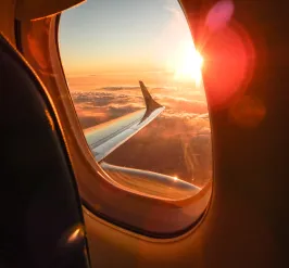 An interior view of an airplane. The plane's wing and a sunset over the clouds can be seen through a window.