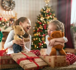 Two sisters open gifts together on Christmas morning. They are both holding stuffed bears.