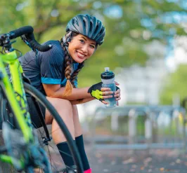 A woman taking a drink from a sports water bottle while taking a break from cycling. She is wearing a bicycle helmet and smiling at the camera.