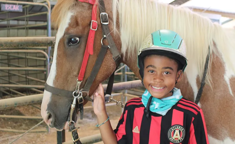 A boy wearing a riding helmet smiles at the camera. He is standing beside a horse.