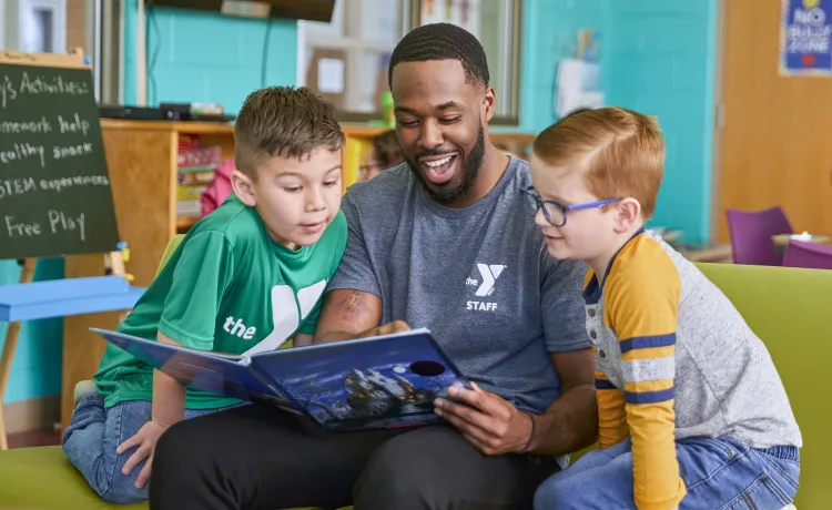 A Y team member reads with two young boys during the YMCA's afterschool program. He is smiling while they look at the open book together.