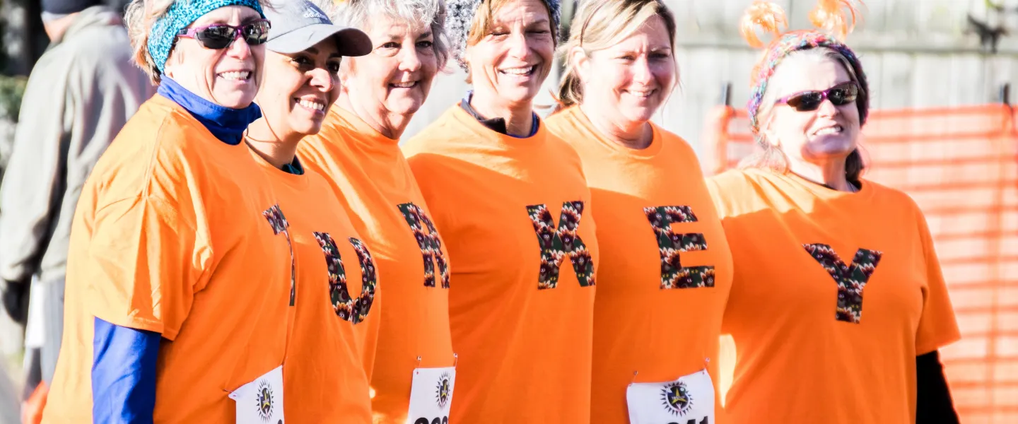 A group of women standing together. They are each wearing orange shirts with a letter on them. The shirts spell out "tirkey."