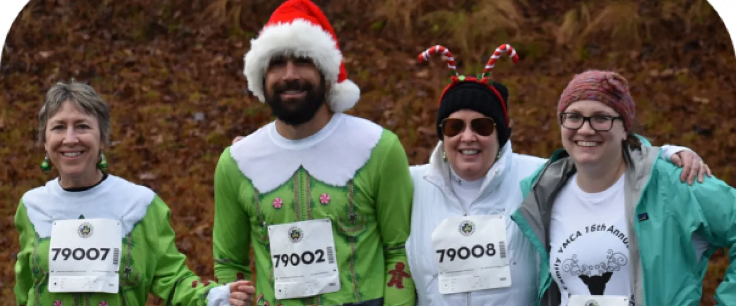 A group of runners in the Ragsdale YMCA's Reindeer Romp standing together. Two of them are wearing green elf costume shirts. A man holds a leash as his dog stands beside him.