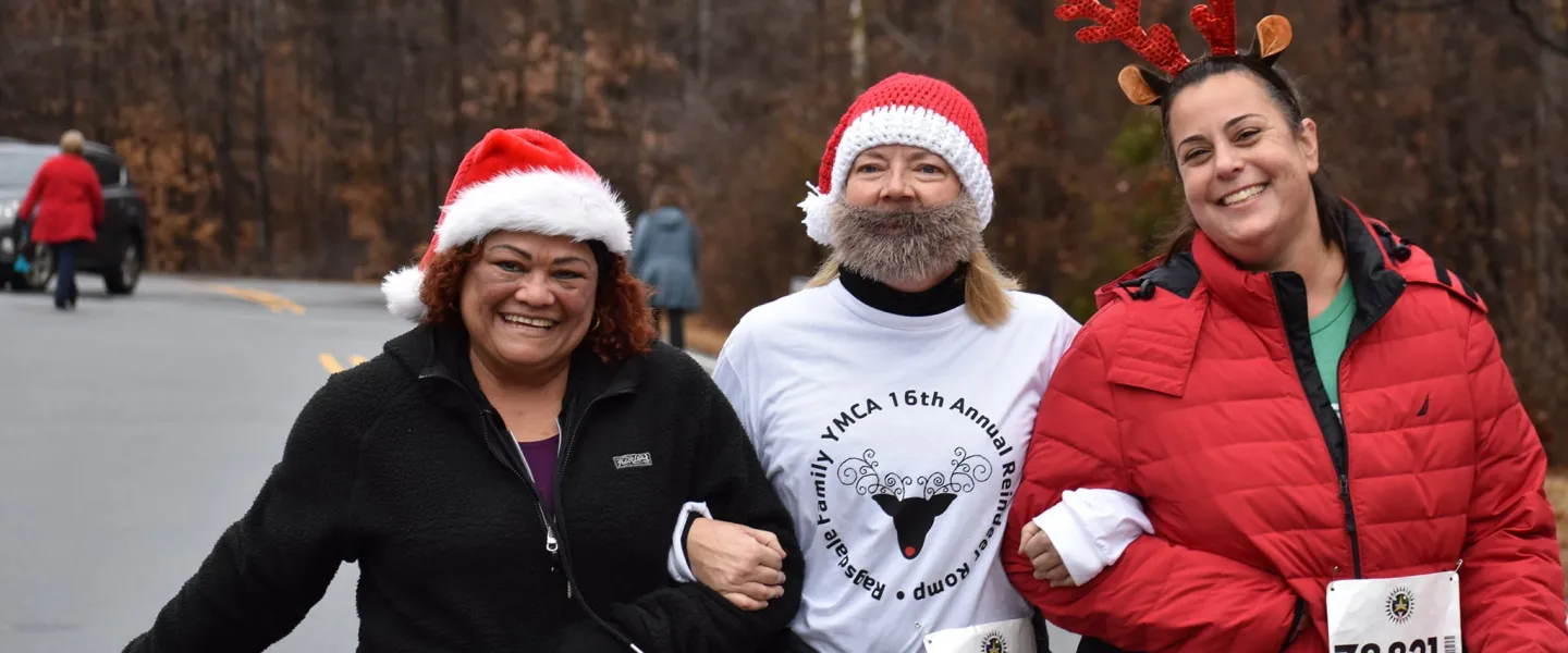 A group of women smiling at the camera. One woman is wearing a Santa hat, while another wears reindeer antlers. Another woman is wearing a Santa hat and beard.