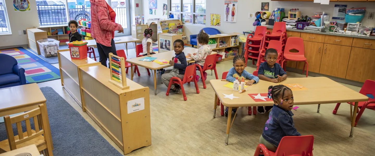 A photo of the Hayes-Taylor YMCA's daycare room. A group of children sit at low tables while a teacher stands nearby and speaks to them.