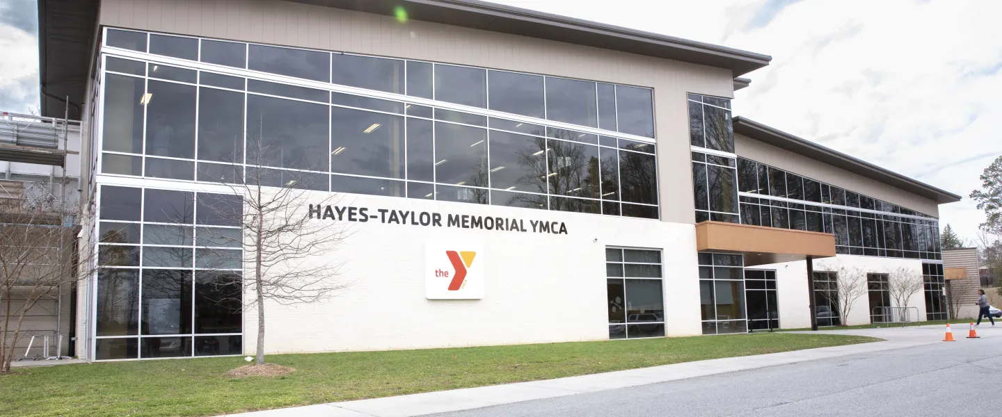 An exterior photo of the Hayes-Taylor YMCA from the front of the branch. There is a sign in black letters that says Hayes-Taylor Memorial YMCA. Below that, there is a red and yellow YMCA logo.
