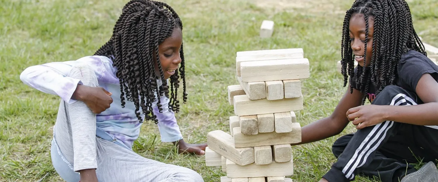 Two children sitting in the grass together and stacking a tower of wooden blocks.