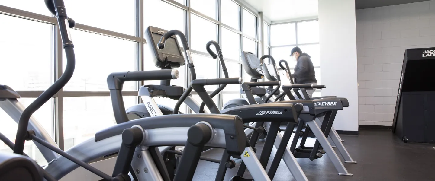 A photo of the wellness center at the Bryan YMCA. There is a line of exercise machines. A woman is working out in the rear of the photo.