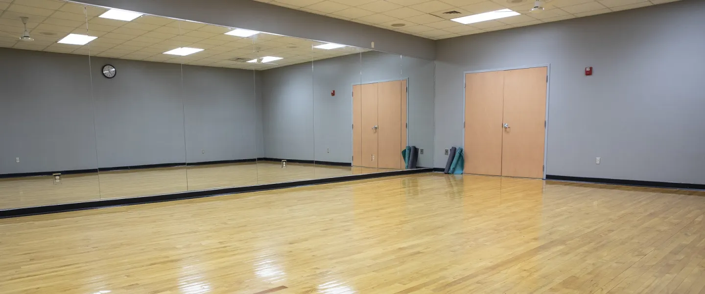 A yoga room at the Bryan YMCA. The wall at the top and left of the photo is covered in a full mirror, and the floor is hardwood. A set of double doors are on the wall at the top-right of the photo.