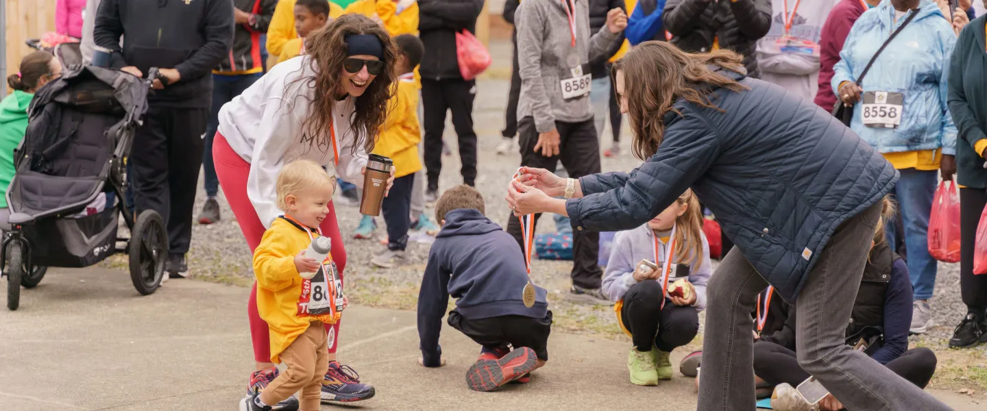 A diverse group of participants at the YMCA of Greensboro Turkey Trot gathered together. Heather Whitsett is approaching a toddler to give them an award medal.