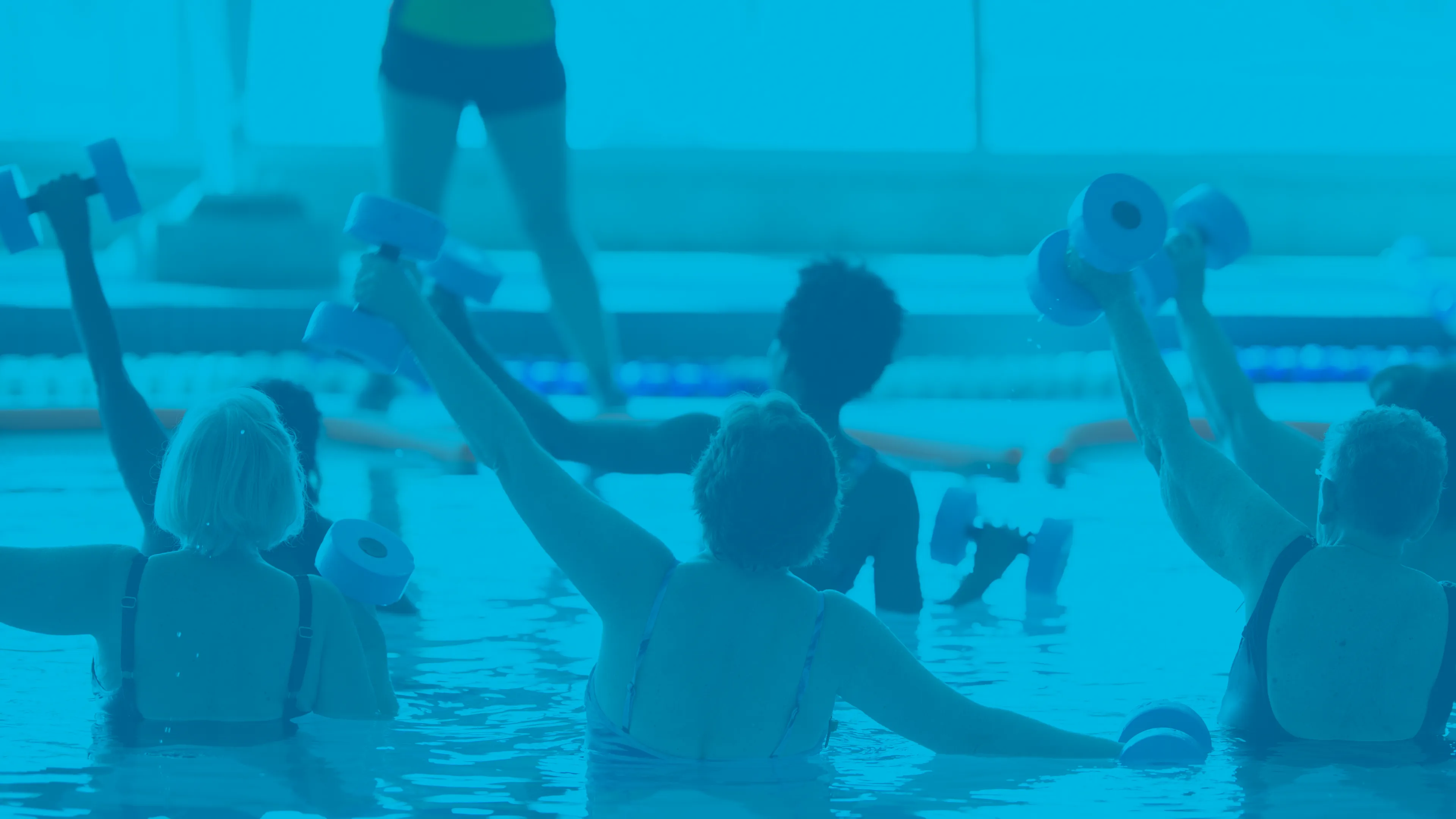 A group of women in a water fitness class. They are exercising with weights and their backs are to the camera while the instructor speaks to them.