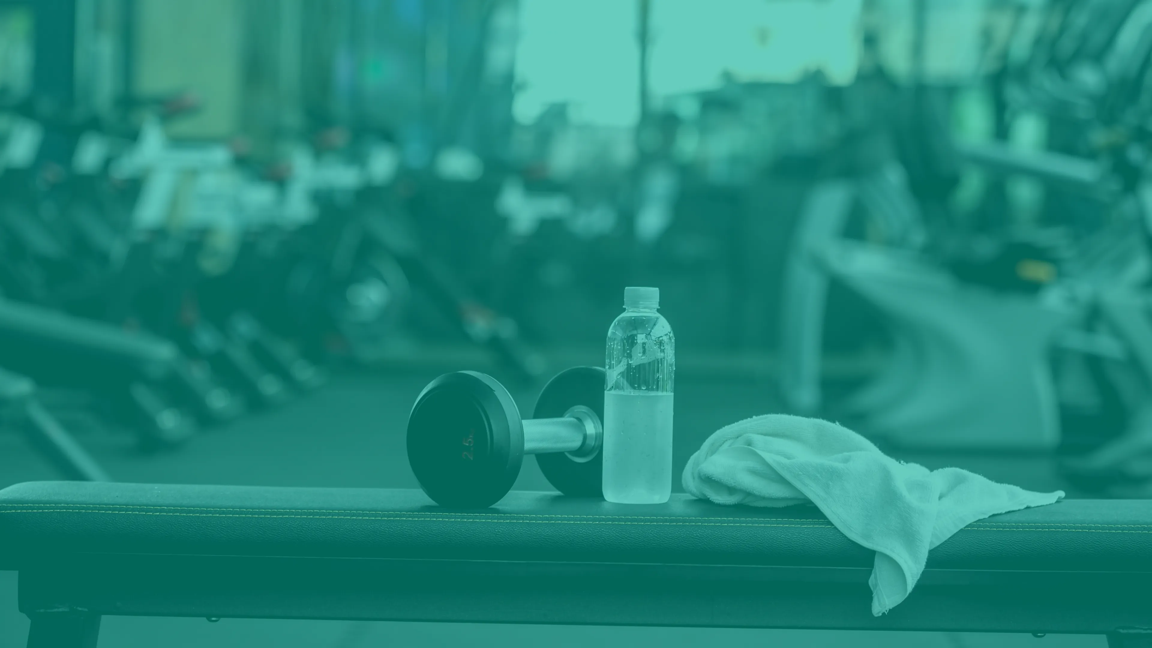 A weight bench in a fitness area. A white towel, a half-filled waterbottle, and a hand weight sit on top of the bench.