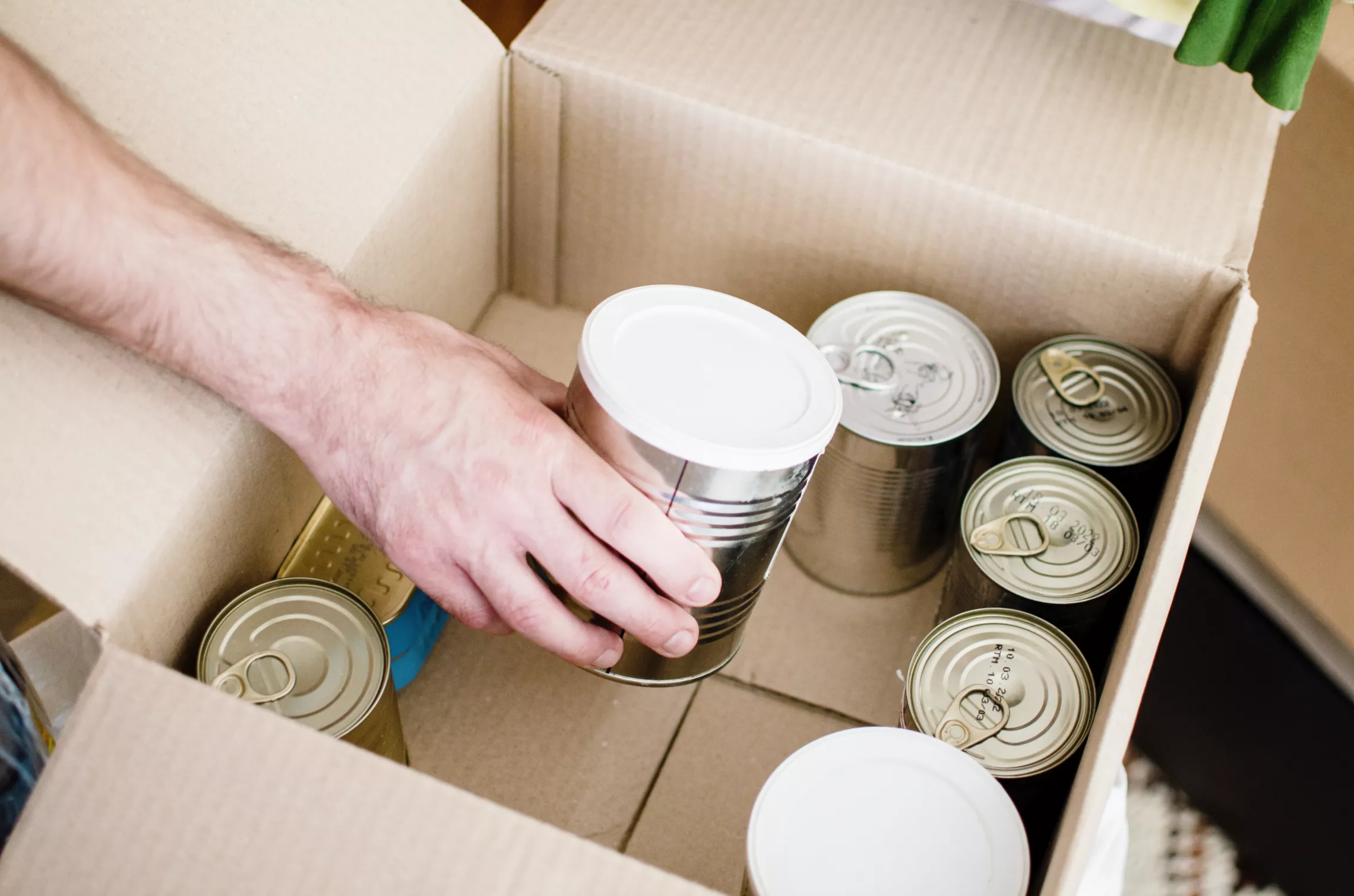 A photo of canned goods inside a cardboard box. There is a man's arm reaching to pick up a can.