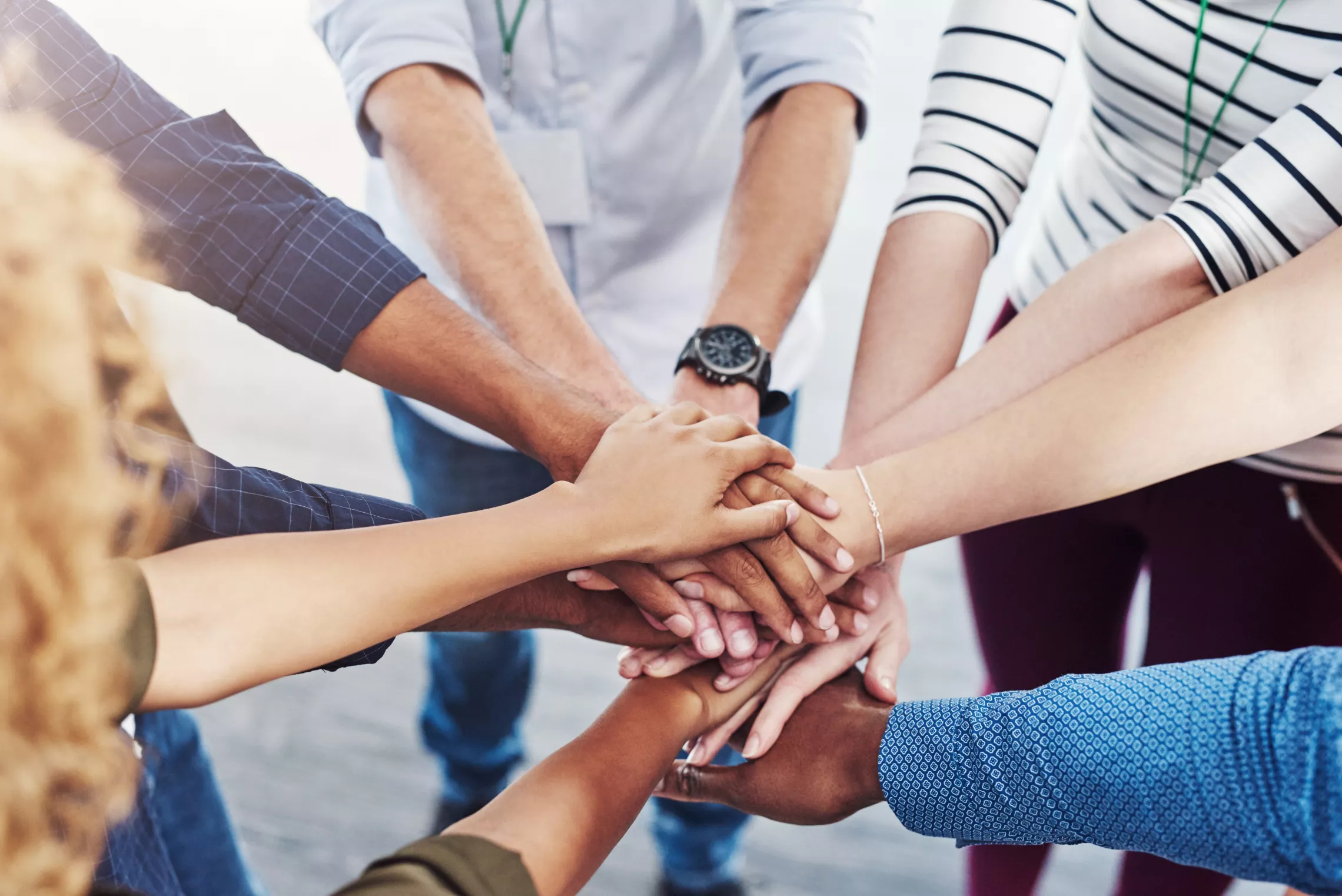 A diverse group of people standing in a circle. Their hands are stacked in the center.