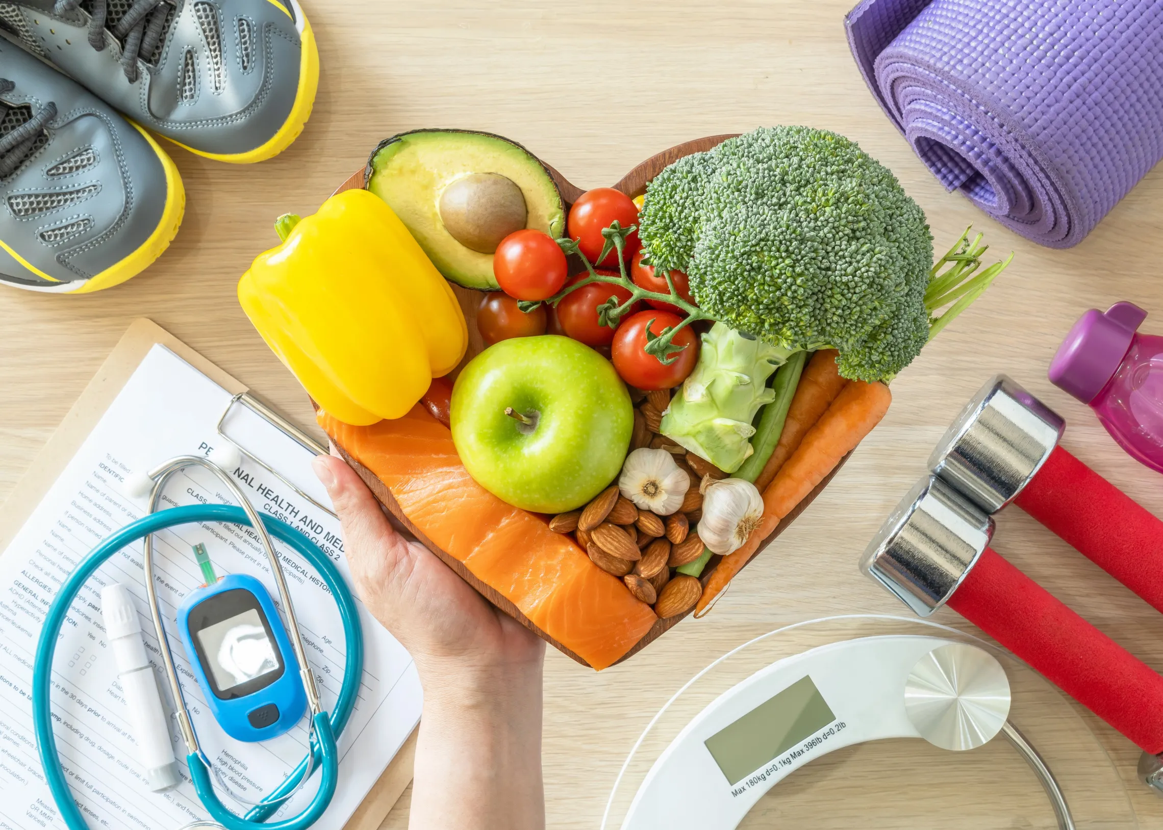 A hand holding a collection of healthy foods. On the table is a collection of fitness equipment, a scale, and other medical devices.