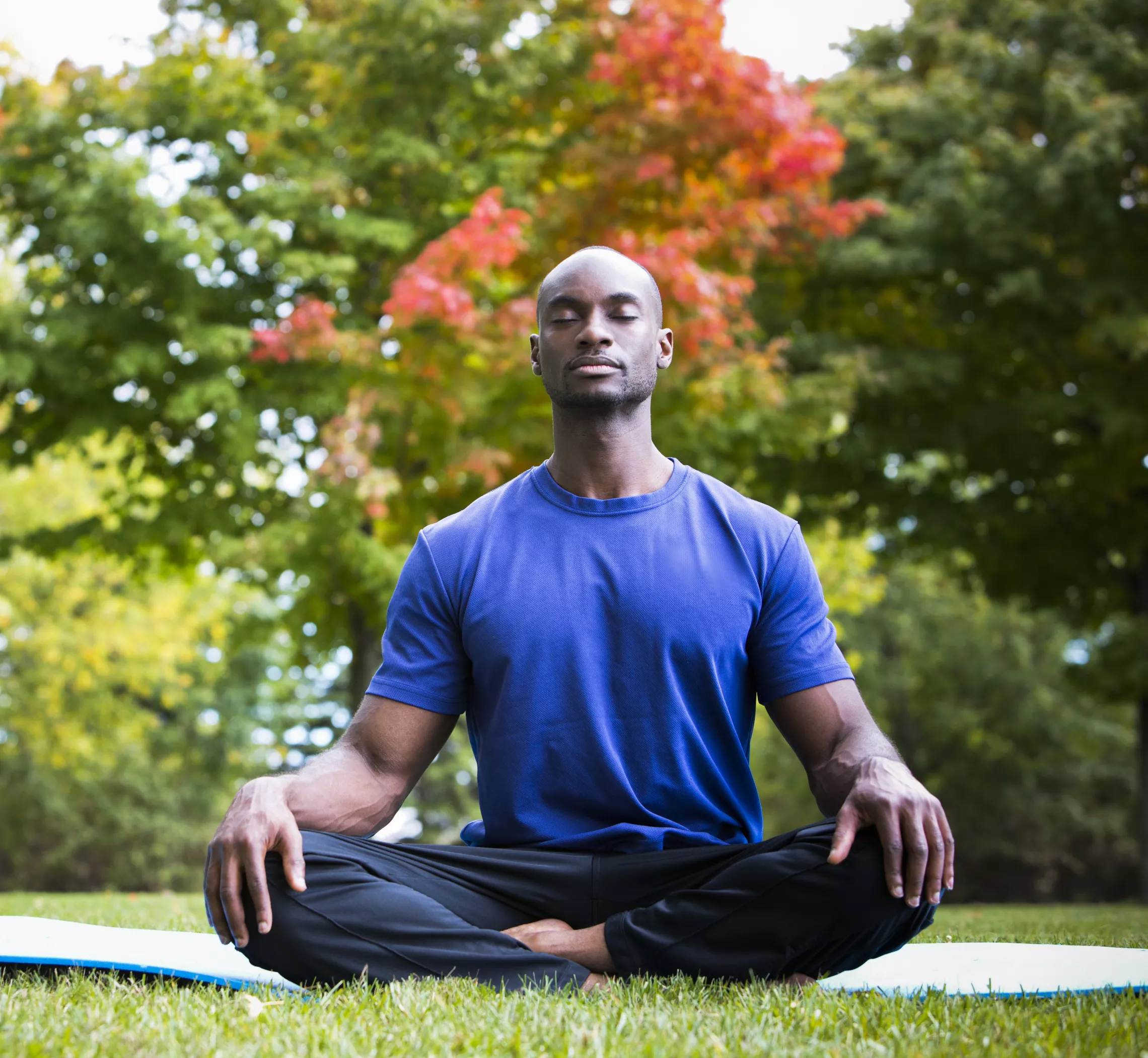 A man sitting in the grass outside on a yoga mat. He has his eyes closed and is sitting cross-legged in a yoga pose.