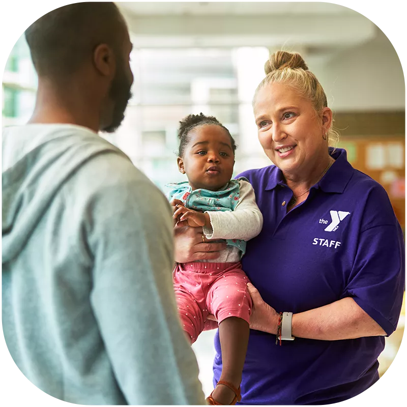 A Y team member holding an infant child in her arms while speaking with their father. The Y team member is smiling.