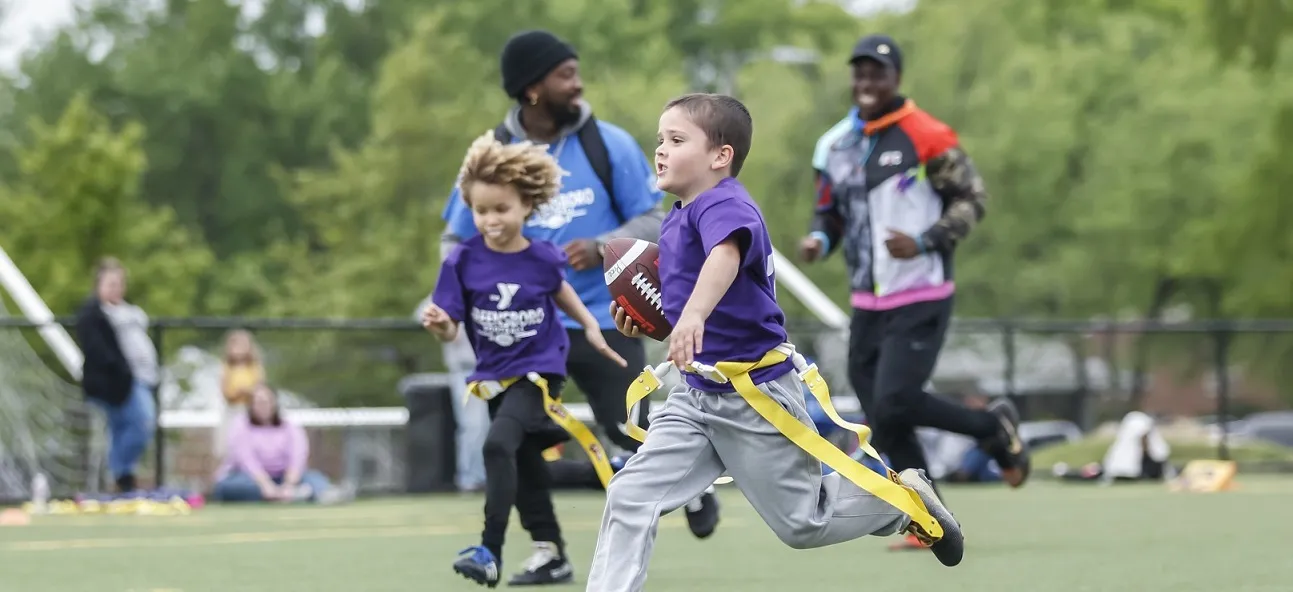 Children playing flag football at the Bryan YMCA. Two Y team members are running alongside a young boy carrying the ball.