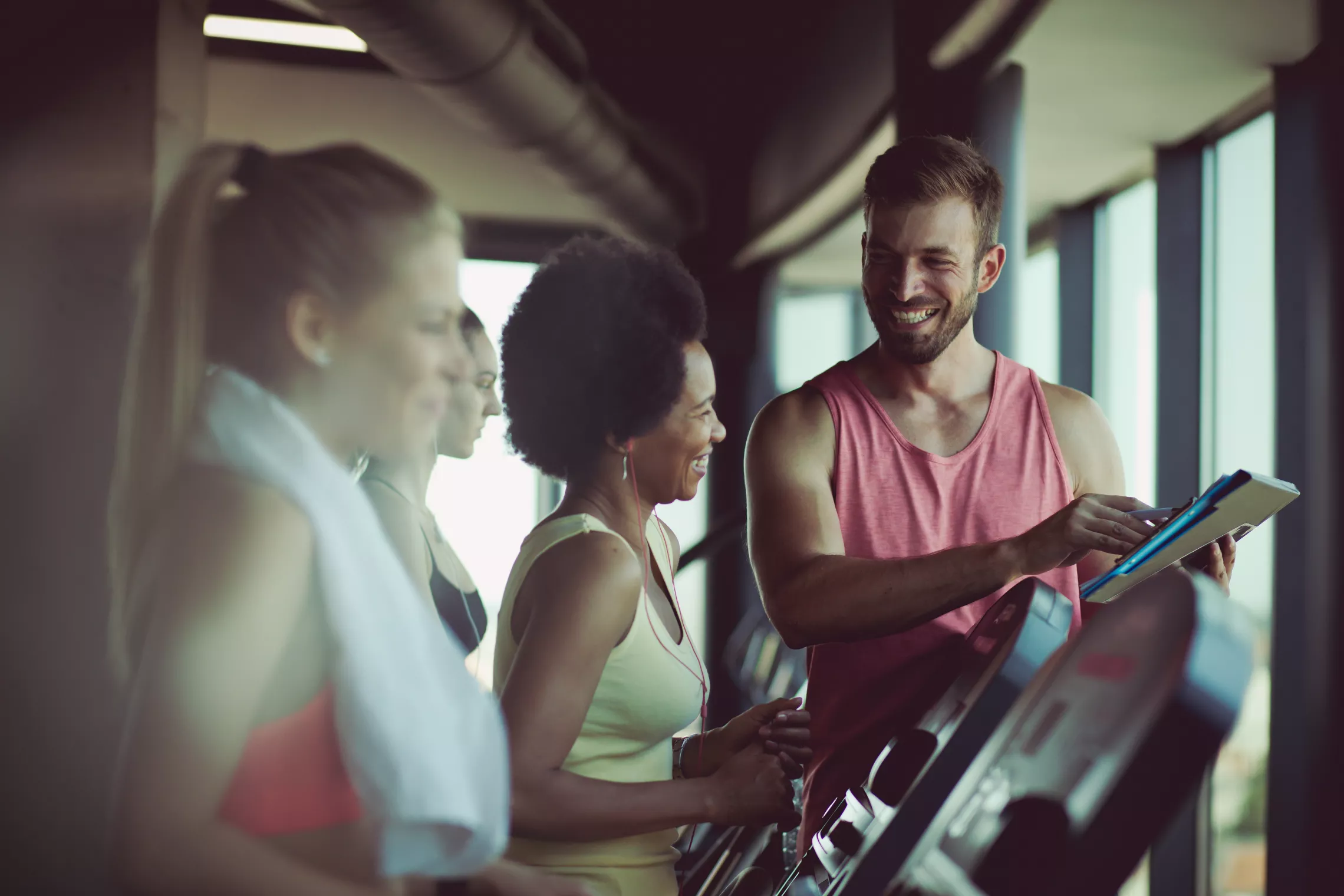 A personal trainer and his client at a gym. She is smiling at him as he assists her while she is using the treadmill.