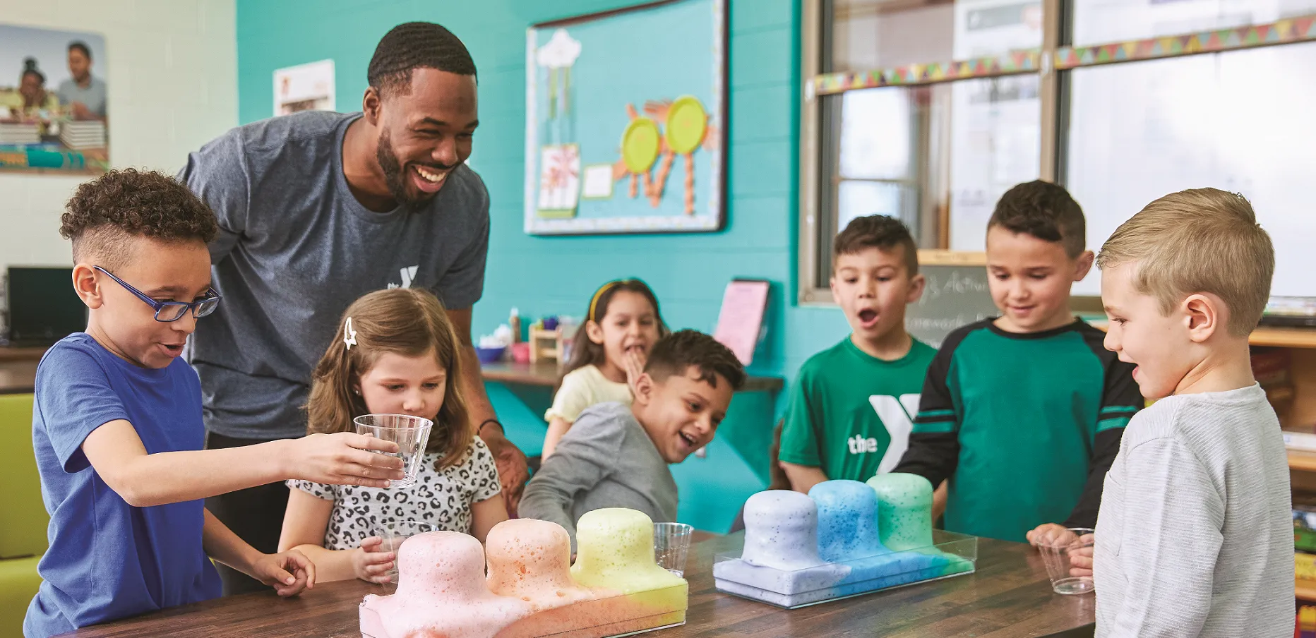 A Y team member stands with a group of children during the Y's Afterschool program. They are all laughing together while doing a science experiment with colorful beakers.