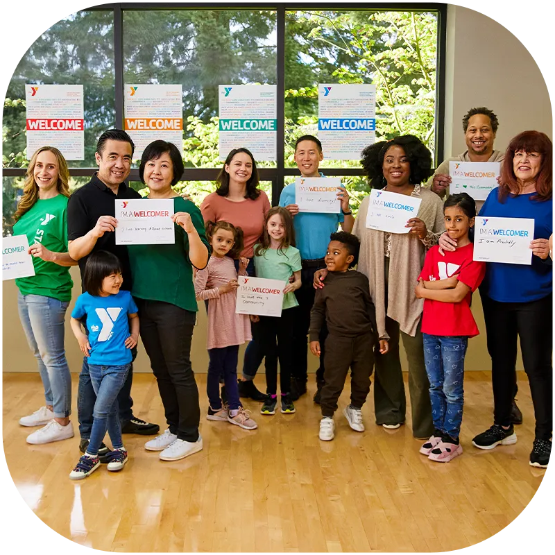 A diverse group of adults and children stand together in a YMCA branch during Welcoming Week.