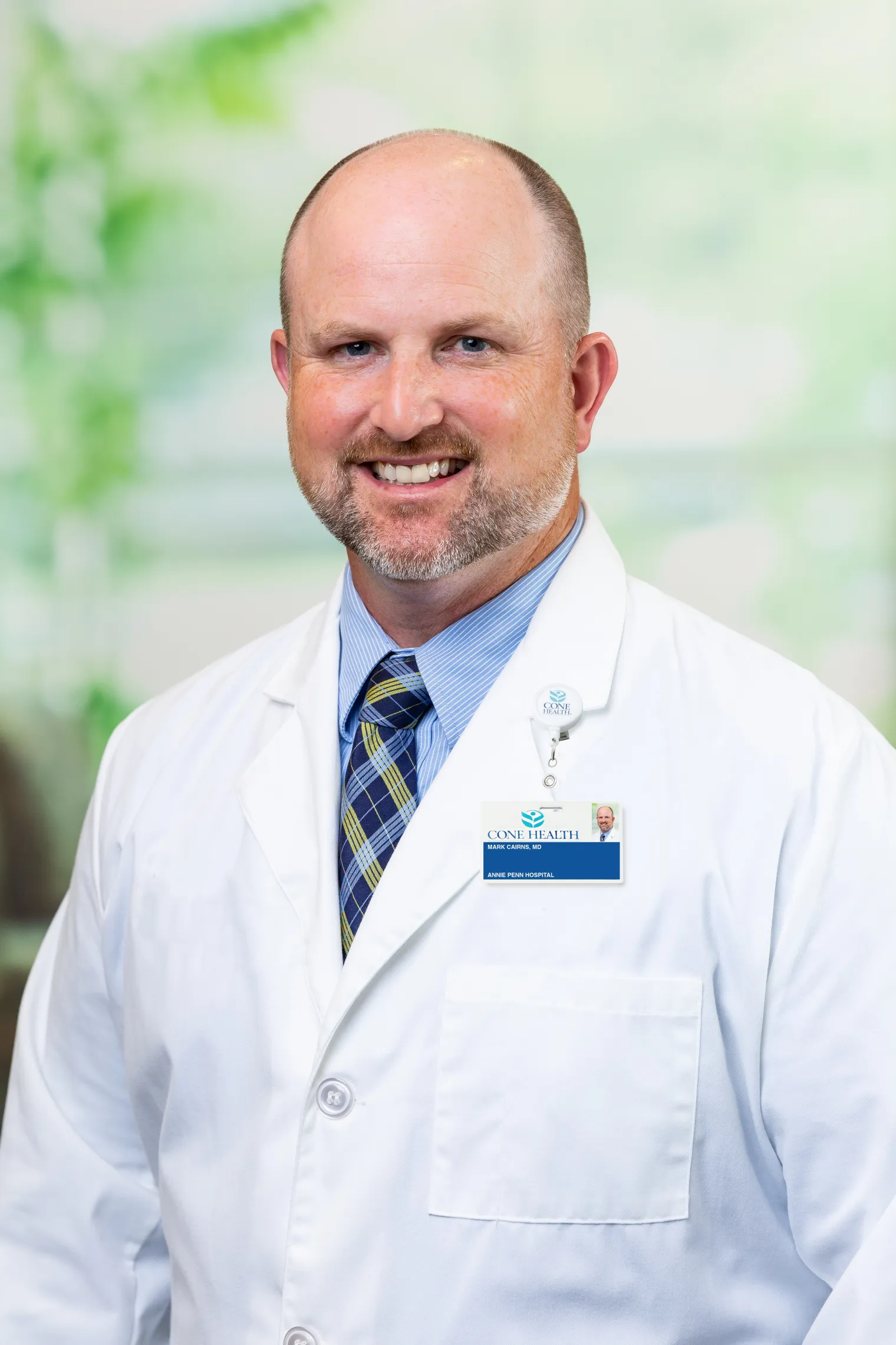 A portrait of Dr. Mark Cairns, MD, MS. A man wearing a white laboratory coat smiles at the camera. He is wearing a blue striped button down shirt with a dark blue tie with yellow and light blue stripes.
