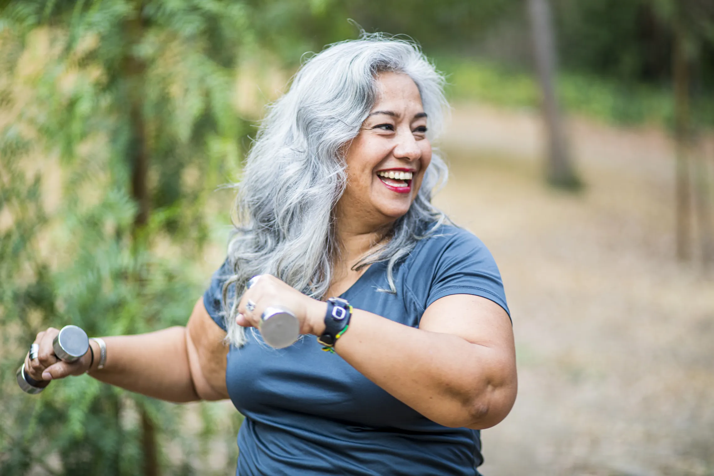 A senior woman smiles while exercising using hand weights outdoors.