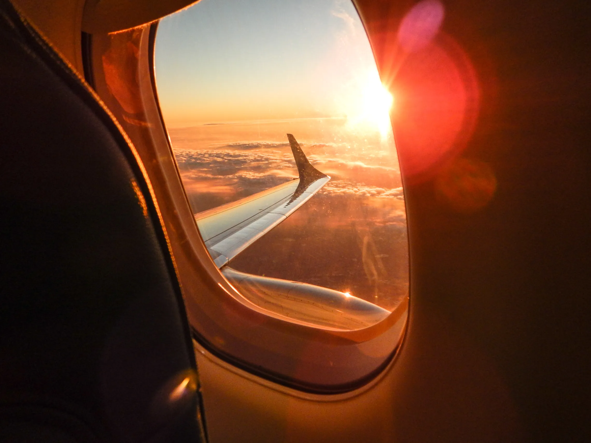 An interior view of an airplane. The plane's wing and a sunset over the clouds can be seen through a window.