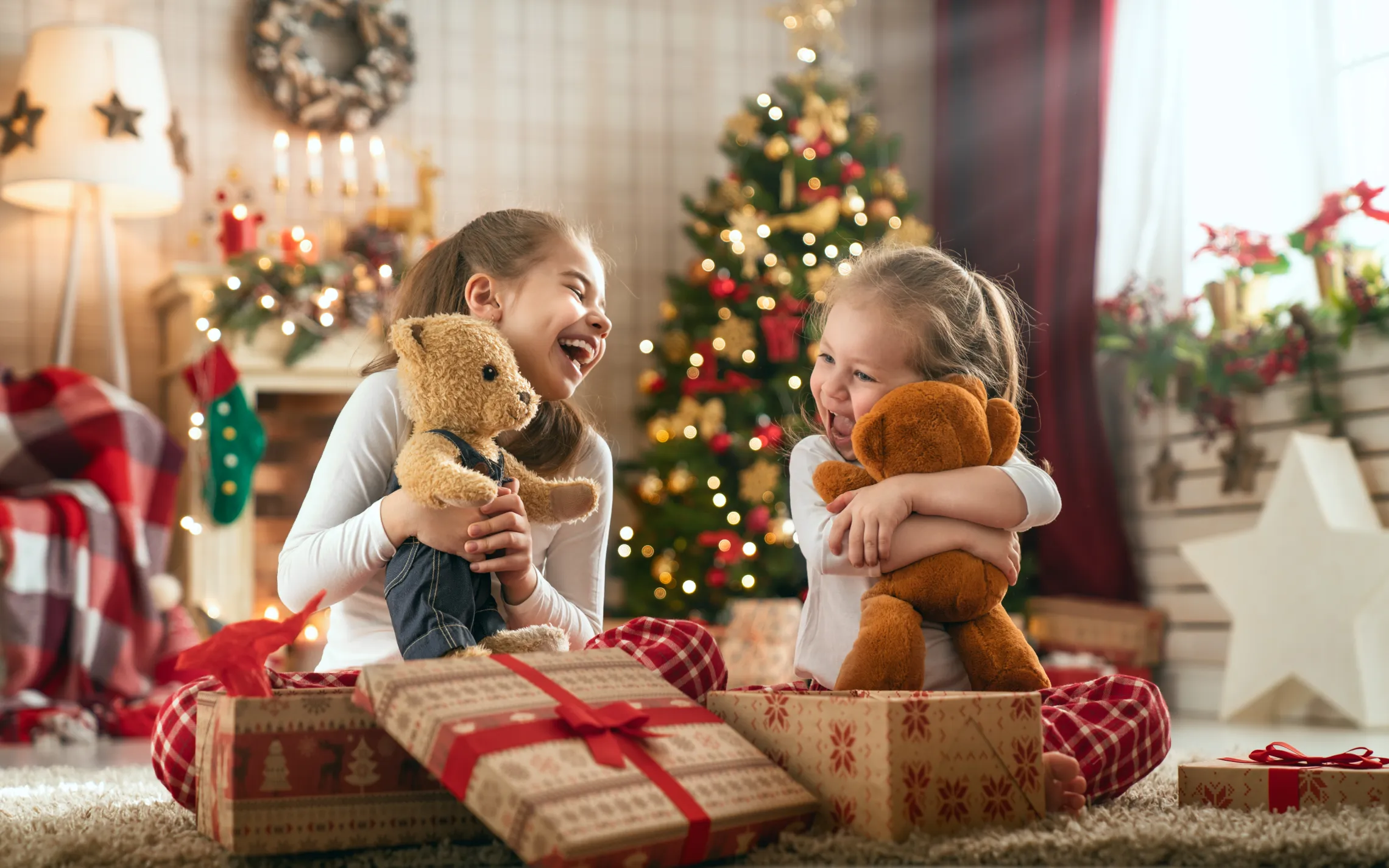 Two sisters open gifts together on Christmas morning. They are both holding stuffed bears.