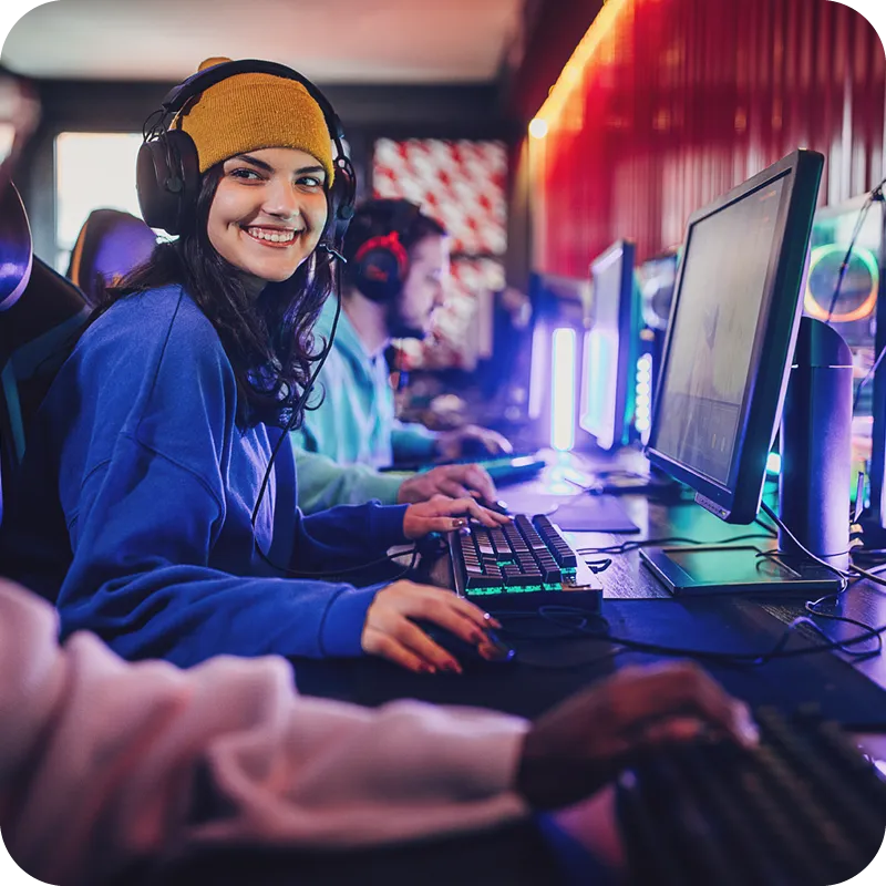 A teenage girl wearing a blue sweatshirt and yellow knit hat smiles at her teammate while playing eSports.