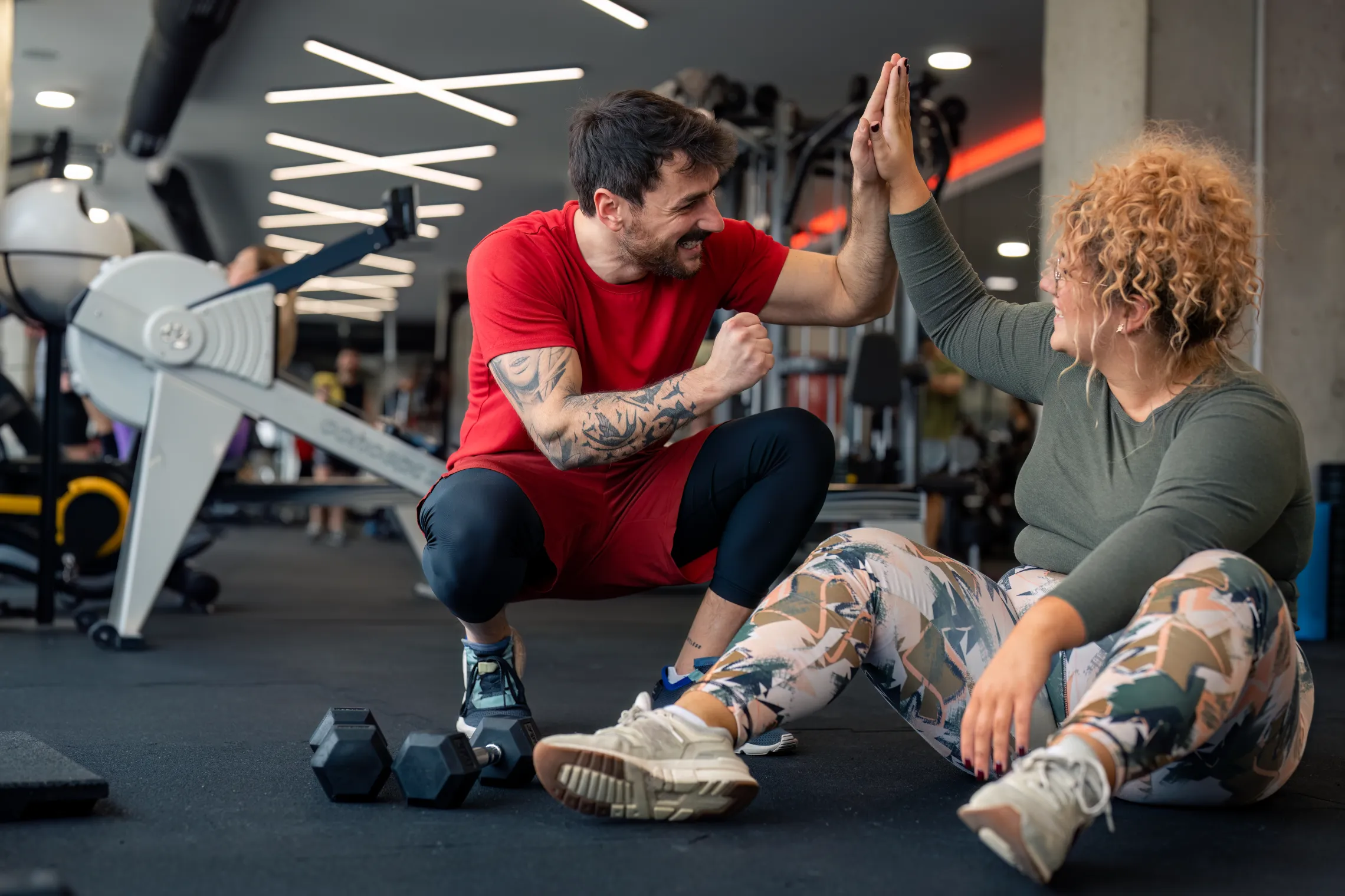 A personal trainer and his client at the gym. The trainer is a man with dark hair and dark facial hair kneeling beside his client, a woman with blond curly hair, sitting on the gym floor. The personal trainer and client are giving each other a high five.