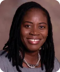 A photo of an African American woman smiling at the camera. She is wearing two strings of pearls and a gray shirt.