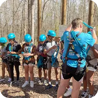 A group of teens in the YMCA of Greensboro Achievers standing together outdoors. They are wearing helmets and equipment for a climbing rope course.