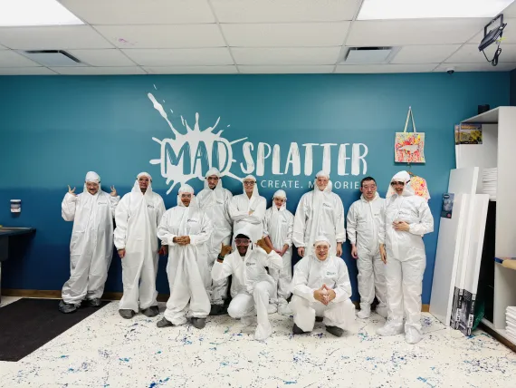 A group photo of QUAD F participants in a room with blue walls and a white floor where paint is splattered. They are wearing white jumpsuits and protective goggles with their hoods pulled up.