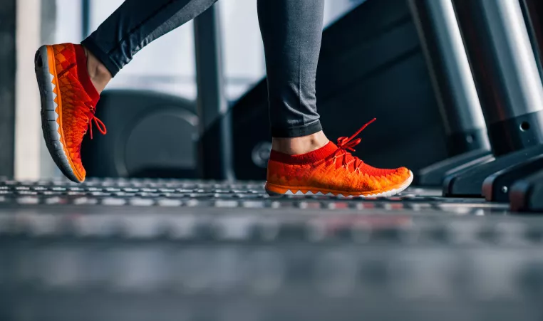 A person wearing gray exercise pants and orange and red shoes walks on a treadmill.