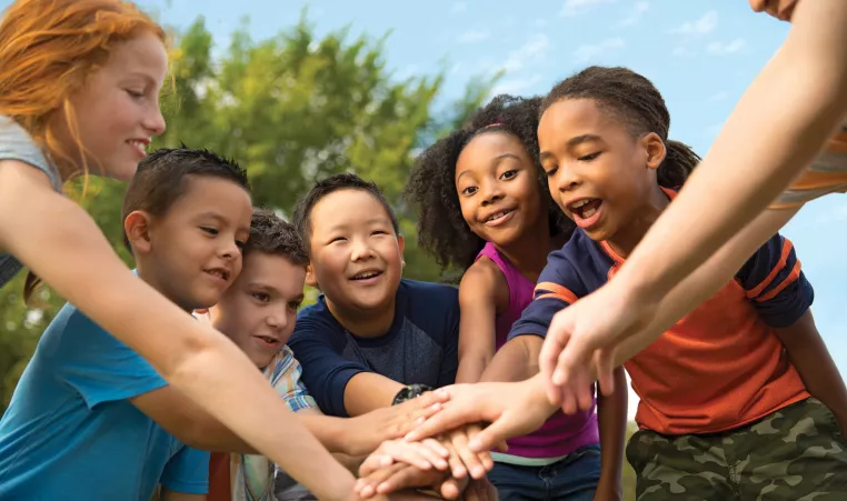 A group of diverse children at summer camp stand in a circle. Their hands are stacked in the middle of the circle.