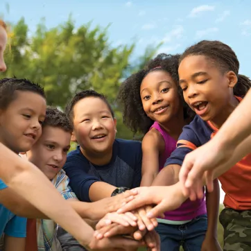 A group of diverse children at summer camp stand in a circle. Their hands are stacked in the middle of the circle.