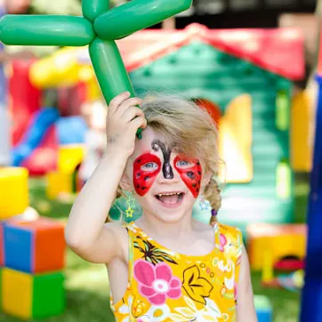 Child with face painting holding a balloon animal in front of bounce house. 