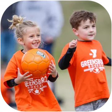 A young boy and girl wearing orange shirts that say Greensboro Soccer in white text. The girl is holding an orange soccer ball and smiling.
