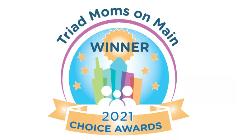 A logo for the Triad Moms on Main Choice Awards. It is a colorful illustration of buildings with white line illustrations of people in front of it. The text around the circle reads "Triad Moms on Main," above the buildings is the word "Winner," then the bottom yellow banner says "2021 Choice Awards" in blue text.