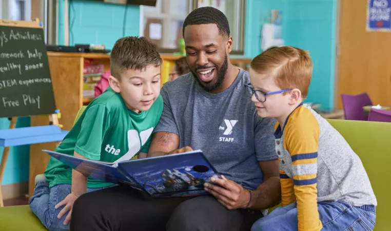 A Y team member reads with two young boys during the YMCA's afterschool program. He is smiling while they look at the open book together.