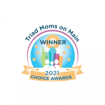 A logo for the Triad Moms on Main Choice Awards. It is a colorful illustration of buildings with white line illustrations of people in front of it. The text around the circle reads "Triad Moms on Main," above the buildings is the word "Winner," then the bottom yellow banner says "2021 Choice Awards" in blue text.