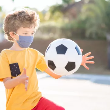 A young boy playing with a soccer ball. He is wearing a mask.