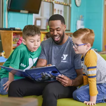 A Y team member reads with two young boys during the YMCA's afterschool program. He is smiling while they look at the open book together.