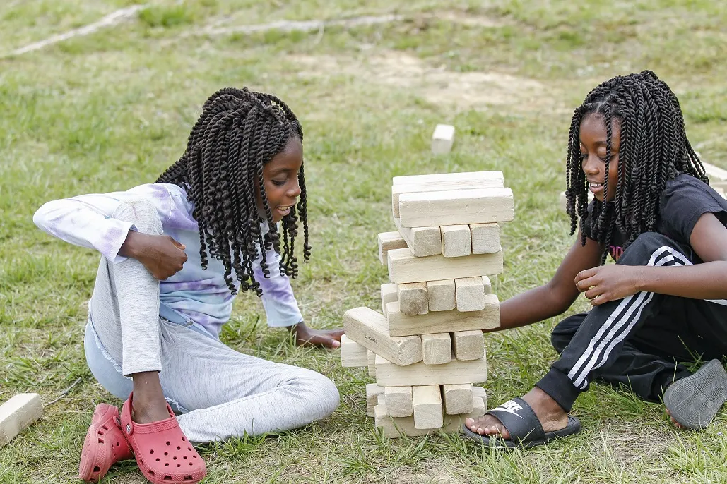 Two children sitting in the grass together and stacking a tower of wooden blocks.