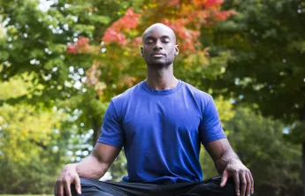 A man sitting in the grass outside on a yoga mat. He has his eyes closed and is sitting cross-legged in a yoga pose.