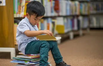 A young boy reading a book. He is sitting on a stack of children's books.